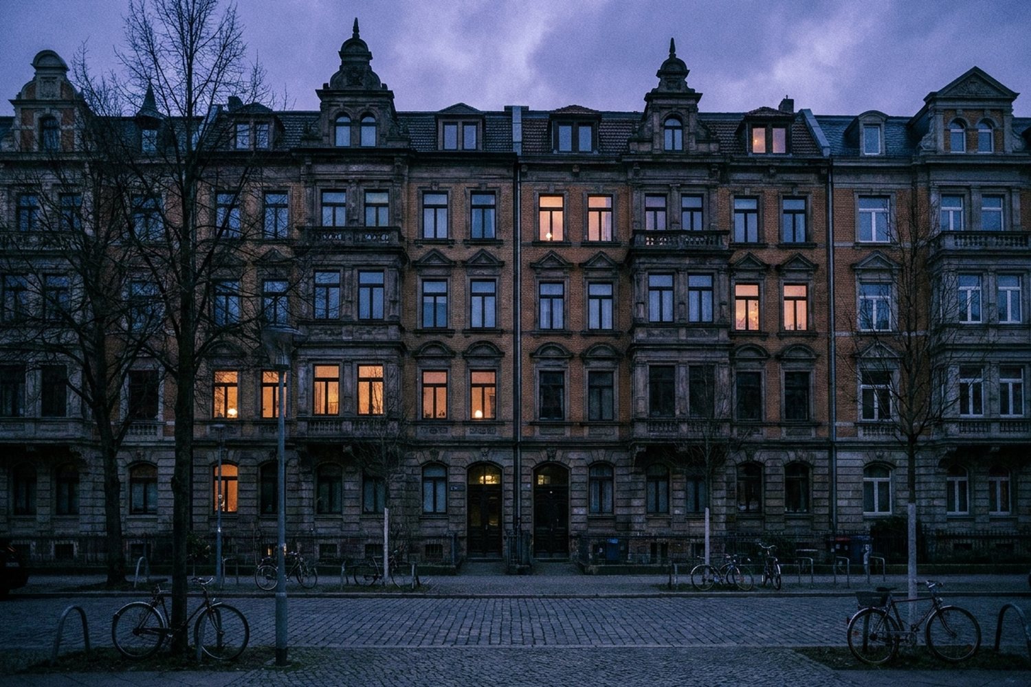 Gründerzeit-Häuserzeile am Abend, hinter vielen Fenstern brennt warmes Licht, davor Fahrräder auf dem Gehweg